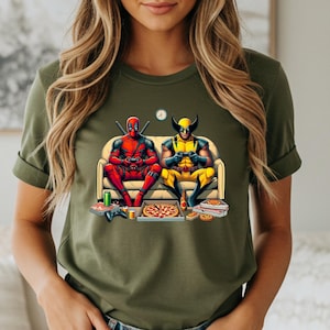 May include: Olive green t-shirt featuring a graphic of Deadpool and Wolverine playing video games on a couch, with pizza and snacks. The characters are in their superhero costumes. A clock is on the wall.