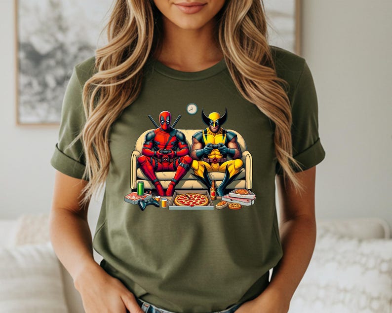 May include: Olive green t-shirt featuring a graphic of Deadpool and Wolverine playing video games on a couch, with pizza and snacks. The characters are in their superhero costumes. A clock is on the wall.