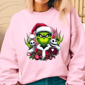 May include: Pink sweatshirt featuring a graphic of the Grinch wearing a Santa hat, with Jack Skellington and a skeleton. The Grinch is green with a white fur trim. The graphic includes candy canes and Christmas ornaments.