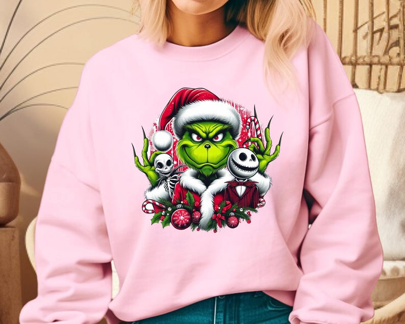 May include: Pink sweatshirt featuring a graphic of the Grinch wearing a Santa hat, with Jack Skellington and a skeleton. The Grinch is green with a white fur trim. The graphic includes candy canes and Christmas ornaments.