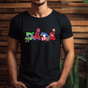 May include: Black t-shirt featuring the word "DAD" with superhero characters. The letter "D" has a green Hulk, the "A" has a Spider-Man, and the second "D" has Iron Man, Captain America, and other characters.