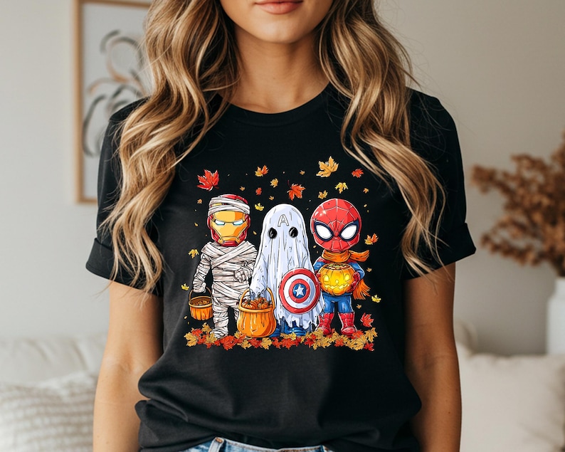 May include: Black t-shirt featuring a Halloween-themed graphic of three costumed characters: Iron Man as a mummy, a ghost with a Captain America shield, and Spider-Man holding a pumpkin. The design includes autumn leaves and pumpkins.