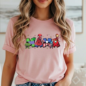 May include: A light pink t-shirt featuring the word "MAMA" in colorful, superhero-themed lettering. Each letter is decorated with a different superhero character, including Hulk, Spider-Man, Captain America, and Iron Man. The shirt has short sleeves.