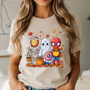 May include: Beige t-shirt featuring a Halloween-themed graphic. The graphic depicts three cartoon characters dressed as Iron Man, a ghost, and Spider-Man, each holding a pumpkin bucket. Autumn leaves surround the characters.