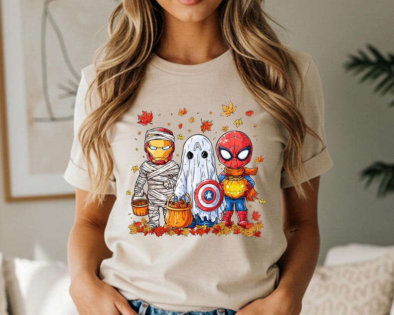 May include: Beige t-shirt featuring a Halloween-themed graphic. The graphic depicts three cartoon characters dressed as Iron Man, a ghost, and Spider-Man, each holding a pumpkin bucket. Autumn leaves surround the characters.