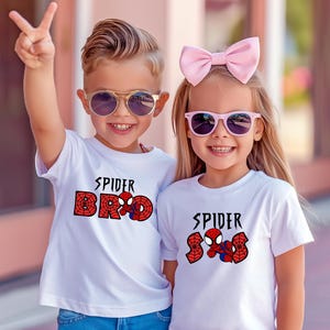 May include: Two children wearing white t-shirts with a Spider-Man graphic. One shirt says "Spider Bro" and the other says "Spider Sis". Both children are wearing sunglasses and smiling. One child is holding up two fingers.