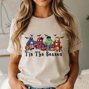 Puede incluir: Camiseta beige con un dise&ntilde;o de cinco fantasmas de superh&eacute;roes con disfraces de Halloween, cada uno sosteniendo una linterna de calabaza. El texto "Tis The Season" est&aacute; impreso debajo, con murci&eacute;lagos.