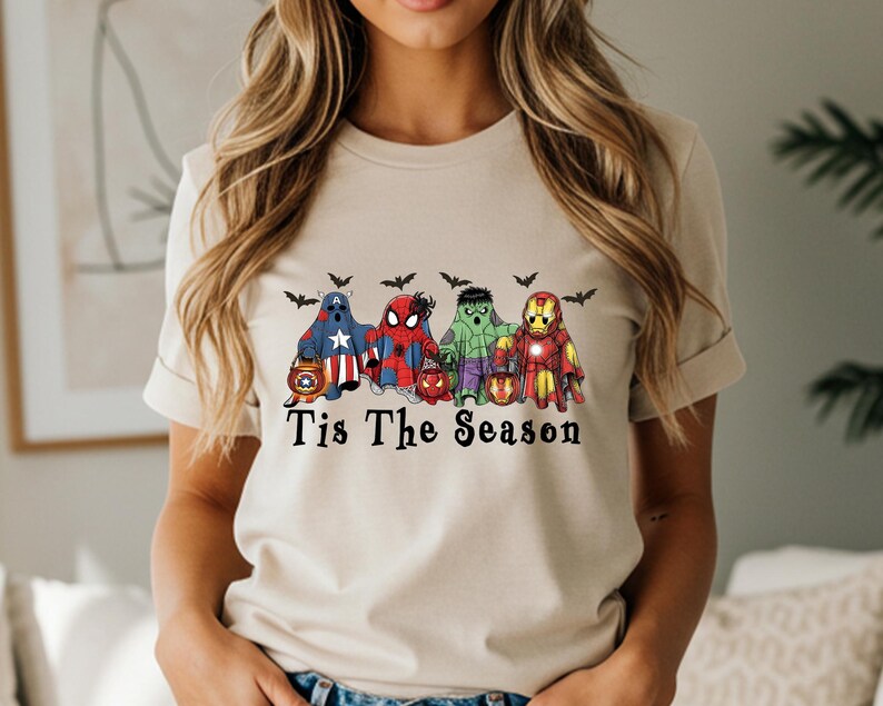 Puede incluir: Camiseta beige con un dise&ntilde;o de cinco fantasmas de superh&eacute;roes con disfraces de Halloween, cada uno sosteniendo una linterna de calabaza. El texto "Tis The Season" est&aacute; impreso debajo, con murci&eacute;lagos.