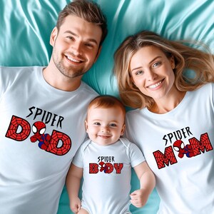 May include: White t-shirts and a onesie with a Spider-Man theme. The shirts say "Spider Dad", "Spider Mom", and "Spider Boy" in black text with a red web design and a Spider-Man graphic. Family matching apparel.