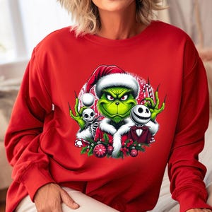 May include: A red long-sleeve sweatshirt featuring a graphic of the Grinch wearing a Santa hat, with Jack Skellington and a skeleton. The Grinch is green with a grumpy expression. The design includes candy canes and Christmas ornaments.