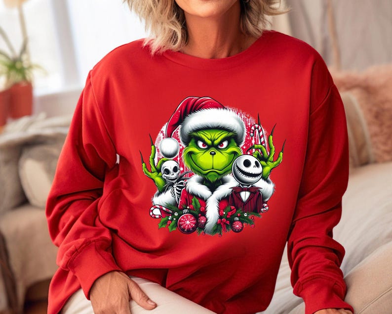 May include: A red long-sleeve sweatshirt featuring a graphic of the Grinch wearing a Santa hat, with Jack Skellington and a skeleton. The Grinch is green with a grumpy expression. The design includes candy canes and Christmas ornaments.