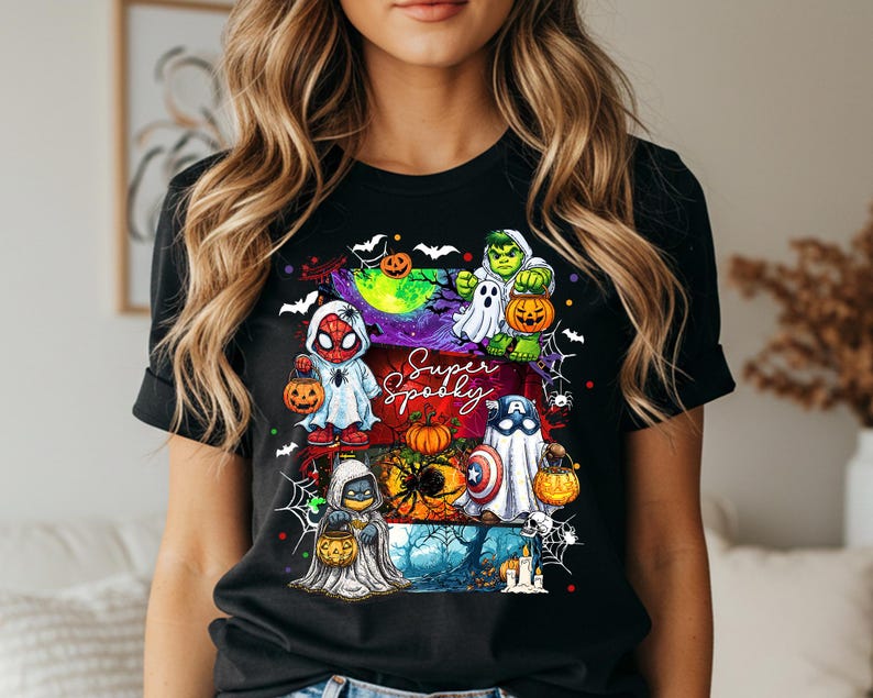 May include: Black t-shirt featuring a colorful Halloween graphic with superheroes dressed as ghosts and holding jack-o'-lanterns. The text "Super Spooky" is written in a script font. The design includes bats, spiderwebs, and a spooky forest scene.