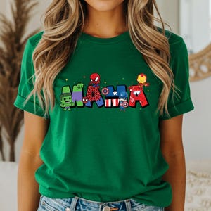 Puede incluir: Una camiseta verde con la palabra "MARVEL" en letras de dibujos animados. Cada letra est&aacute; dise&ntilde;ada con un personaje diferente de Marvel, incluyendo Hulk, Spider-Man, Capit&aacute;n Am&eacute;rica e Iron Man. La camiseta tiene mangas enrolladas.