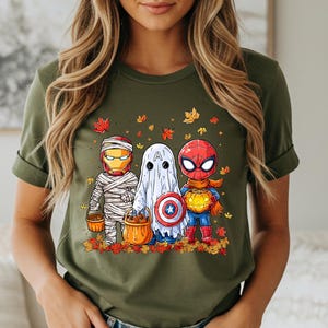 May include: Olive green t-shirt featuring a Halloween-themed graphic. The graphic depicts three costumed characters: Iron Man as a mummy, a ghost, and Spider-Man, all holding pumpkin buckets. Autumn leaves surround the characters.