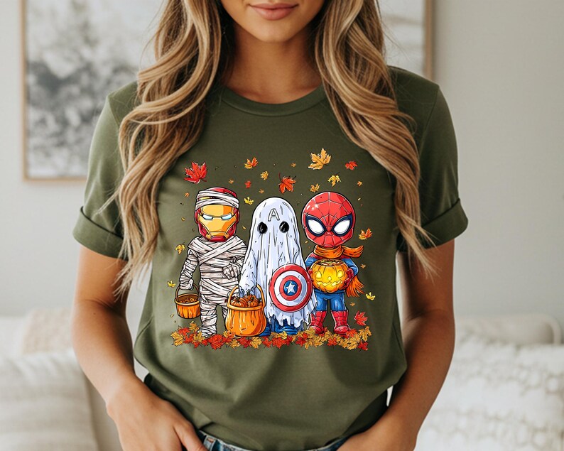 May include: Olive green t-shirt featuring a Halloween-themed graphic. The graphic depicts three costumed characters: Iron Man as a mummy, a ghost, and Spider-Man, all holding pumpkin buckets. Autumn leaves surround the characters.