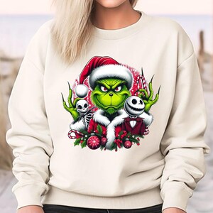 May include: Cream-colored sweatshirt featuring a graphic of the Grinch in a Santa hat, with Jack Skellington and a skeleton. The Grinch is green with a red and white Santa hat. The design includes candy canes and Christmas ornaments.