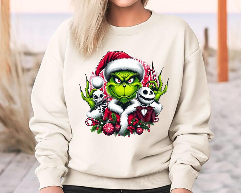 May include: Cream-colored sweatshirt featuring a graphic of the Grinch in a Santa hat, with Jack Skellington and a skeleton. The Grinch is green with a red and white Santa hat. The design includes candy canes and Christmas ornaments.