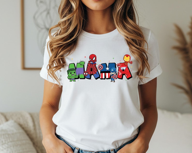 May include: White t-shirt with the word 'MAMA' in colorful superhero-themed letters. The letters are designed with characters like Hulk, Spider-Man, Captain America, and Iron Man. The shirt is a casual wear item, perfect for fans of Marvel comics and superheroes.