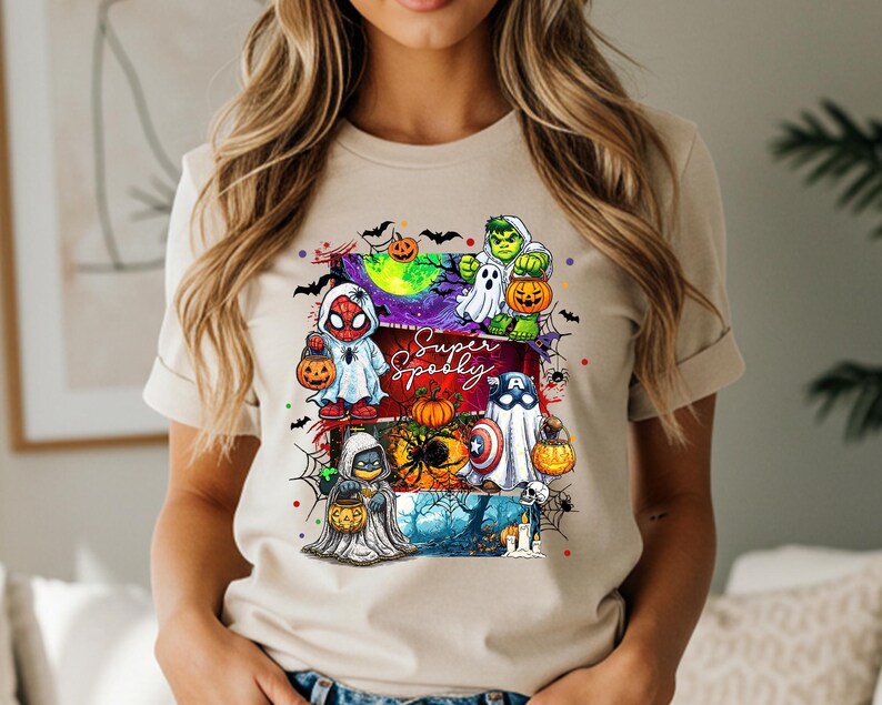 May include: Beige t-shirt featuring a colorful Halloween-themed graphic. The design includes cartoon characters dressed as superheroes, holding jack-o'-lanterns, with the words "Super Spooky" in a stylized font. Bats and spiderwebs complete the festive look.