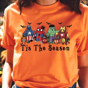 Puede incluir: Camiseta naranja con un dise&ntilde;o de Halloween. Presenta fantasmas de superh&eacute;roes sosteniendo linternas, con murci&eacute;lagos encima. El texto "Tis The Season" est&aacute; impreso debajo. Un dise&ntilde;o divertido y festivo.