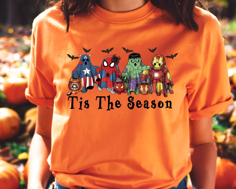 Puede incluir: Camiseta naranja con un dise&ntilde;o de Halloween. Presenta fantasmas de superh&eacute;roes sosteniendo linternas, con murci&eacute;lagos encima. El texto "Tis The Season" est&aacute; impreso debajo. Un dise&ntilde;o divertido y festivo.