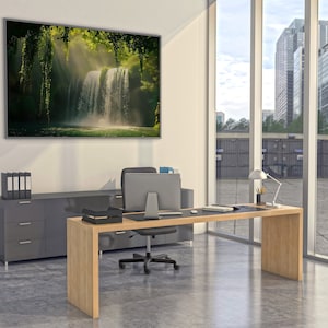 May include: A modern office with a large desk made of light wood. The desk has a black leather top and a black office chair. There is a large window behind the desk with a view of a city skyline. A framed picture of a waterfall in a lush green forest hangs on the wall.