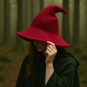 May include: A vibrant red witch hat with a wide brim and a pointed tip. The hat is made of a textured material. The person in the image is wearing a dark green cloak. The background is a blurred forest scene.
