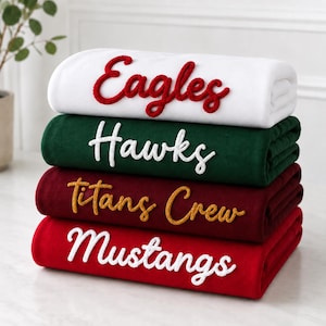 May include: A stack of four folded fleece blankets in white, dark green, maroon, and red. The white blanket features the red embroidered text "Eagles", the green has white "Hawks", the maroon has yellow "Titans Crew", and the red has white "Mustangs".