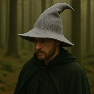 May include: A gray, pointed wizard hat with a wide brim. The hat is made of a knitted material and has a curved tip. The person in the image is wearing a dark green hooded cloak. The background is a blurred forest.