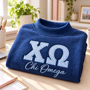 May include: A navy blue sweatshirt featuring a high ribbed collar and long sleeves. The front displays the Greek letters Chi and Omega in white and light blue vertical stripes, with the text Chi Omega embroidered in light blue script below.