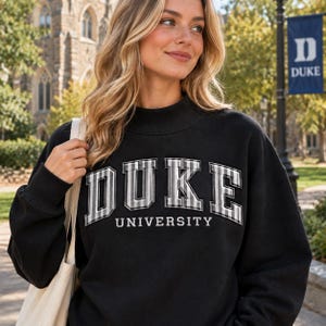 Può includere: Una felpa nera a maniche lunghe con collo alto. La parte anteriore presenta la parola DUKE in grandi lettere in stile blocco con un motivo a quadri grigio e bianco, e la parola UNIVERSITY in caratteri bianchi più piccoli sotto la grafica.