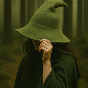 May include: A green witch hat with a wide brim and a pointed tip. The hat is made of a textured fabric and is worn with a matching green cloak. The background is a blurred forest scene.
