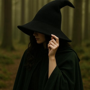 May include: A black witch hat with a wide brim and pointed crown. The hat is worn with a dark green cloak. The person is in a forest setting with blurred trees in the background. The hat is being held by a hand.