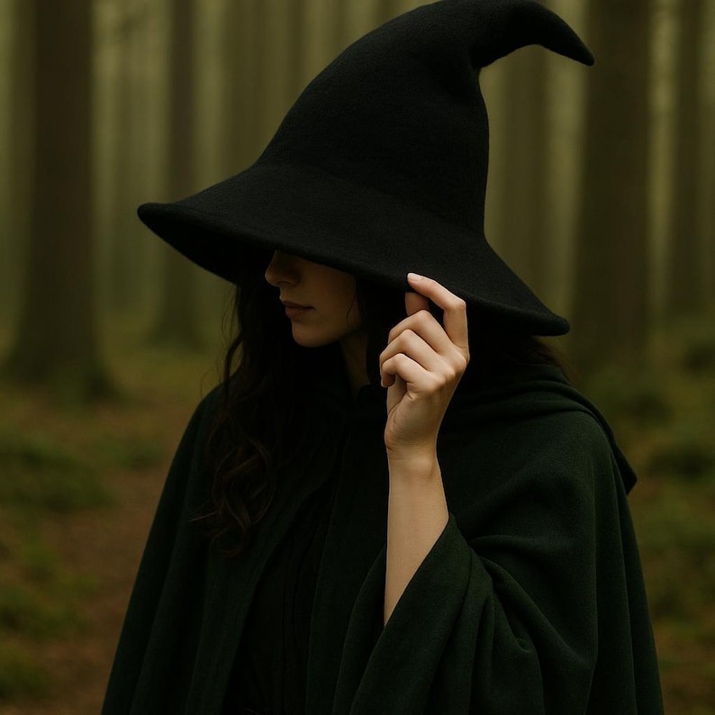 May include: A black witch hat with a wide brim and pointed crown. The hat is worn with a dark green cloak. The person is in a forest setting with blurred trees in the background. The hat is being held by a hand.