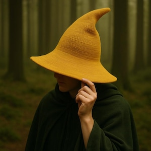 May include: A mustard yellow witch hat with a wide brim and pointed tip. The hat is made of a textured fabric. The person in the image is wearing a dark green cloak. The background is a blurred forest.