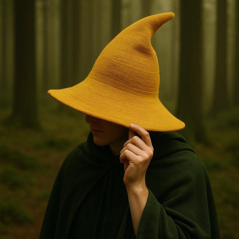 May include: A mustard yellow witch hat with a wide brim and pointed tip. The hat is made of a textured fabric. The person in the image is wearing a dark green cloak. The background is a blurred forest.