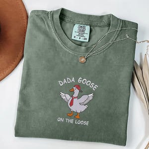 May include: An olive green t-shirt featuring the embroidered text "DADA GOOSE ON THE LOOSE" and a cartoon goose wearing a red hat and tie. A silver necklace with a pendant is draped on the shirt. A brown hat is visible in the upper left corner.