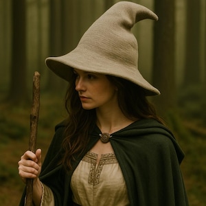 May include: A woman wearing a beige witch hat with a pointed crown and wide brim, a dark green cloak, and a light-colored dress with gold embroidery. She is holding a wooden staff in a forest setting.