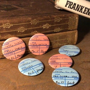 May include: Five vintage-style library card buttons with blue and pink borders. The buttons feature the titles and authors of classic books, including "Little Women", "Frankenstein", "Dracula", "Pride and Prejudice", and "The Lion, the Witch, and the Wardrobe".