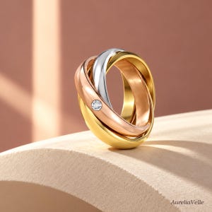 May include: A tri-color interlocking ring featuring three smooth bands in yellow gold, rose gold, and silver tones. The rose gold band is accented with a single small, round, clear gemstone set flush into the metal surface. The brand name AureliaVelle is visible.