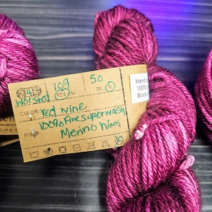 Puede incluir: Un ovillo de hilo de lana merino superwash 100% de color vino tinto. La etiqueta del hilo dice "vino tinto", "100% Fine Superwash Merino Wool" e incluye instrucciones de cuidado.