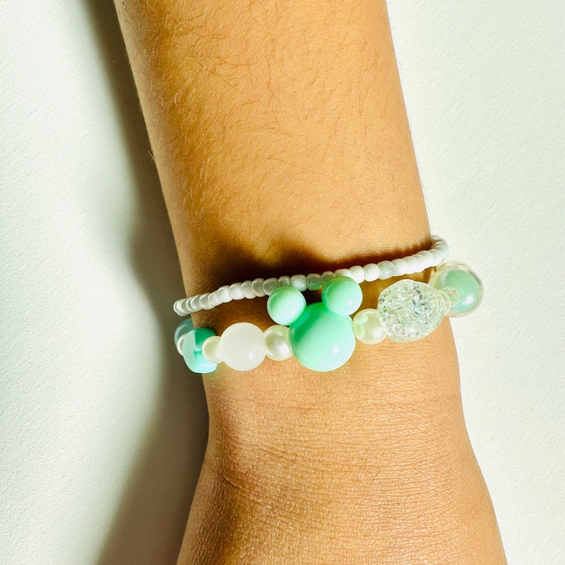 May include: A white and light green beaded bracelet with a white and silver charm. The bracelet has a Mickey Mouse head charm.