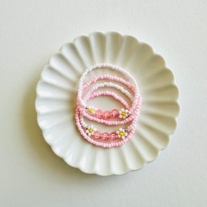 May include: Two pink and white beaded bracelets with the word "BFF" spelled out in white letters on pink beads. Each bracelet has small white flowers with yellow centres.