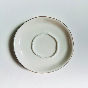 May include: A white beaded bracelet on a white ceramic plate with a gold rim.
