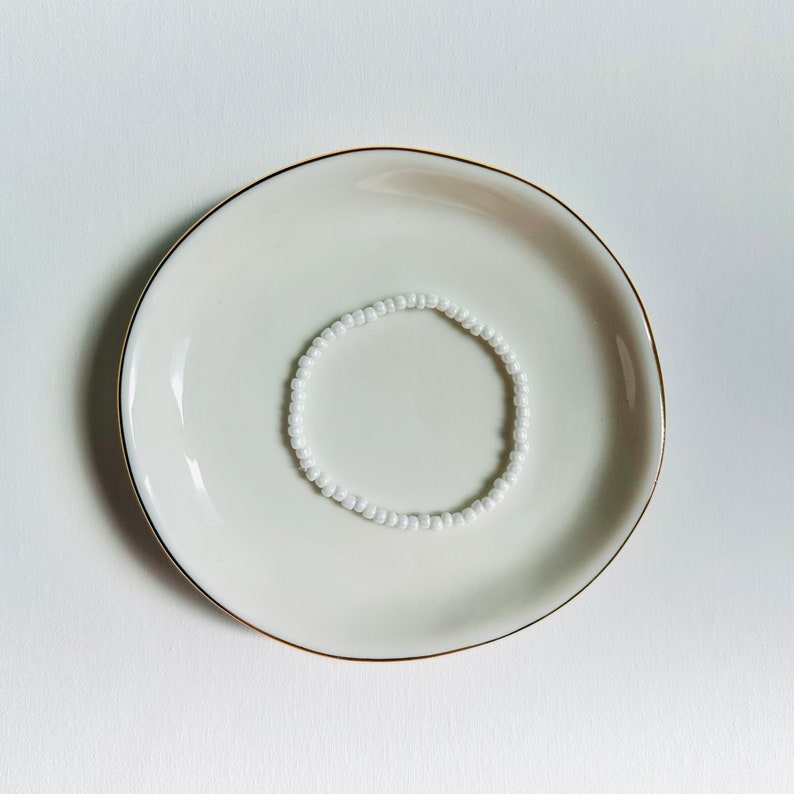 May include: A white beaded bracelet on a white ceramic plate with a gold rim.