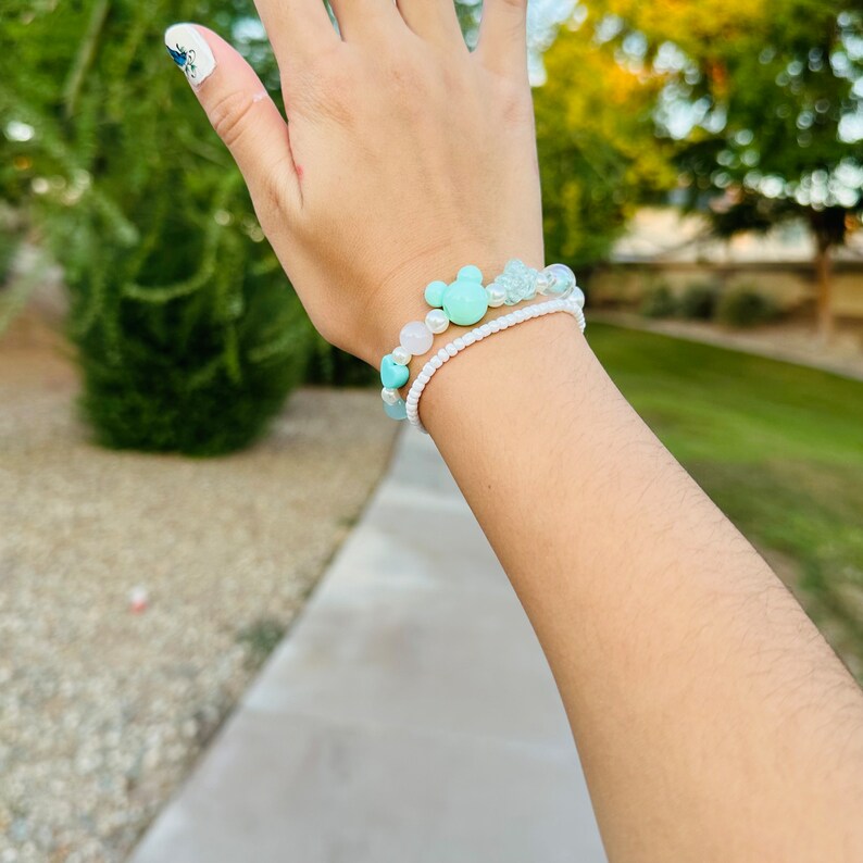 May include: A close-up of a wrist wearing two bracelets. The top bracelet is made of white beads and has a light blue Mickey Mouse charm. The bottom bracelet is made of light blue and white beads.