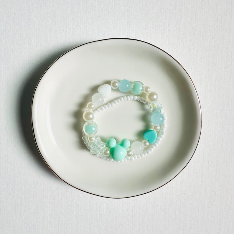 May include: A white and light blue beaded bracelet with a light blue heart charm and a light blue Mickey Mouse charm.