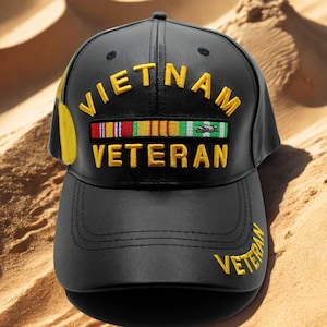 Puede incluir: Gorra de béisbol negra con la inscripción "VIETNAM VETERAN" bordada en amarillo. La gorra presenta una fila de cintas de colores y la palabra "VETERAN" bordada en la visera. La gorra es de material similar al cuero.