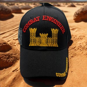 May include: Black baseball cap with "COMBAT ENGINEER" embroidered in red and gold. The cap features a gold castle emblem and additional text on the side. The hat is set against a desert backdrop.