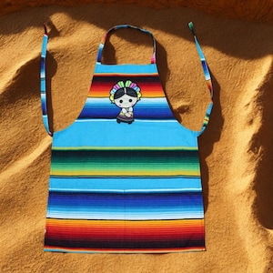 May include: A vibrant apron with a striped pattern in blue, green, red, and yellow. It features a cartoon doll in the centre. The apron has blue straps and is suitable for cooking or crafting.